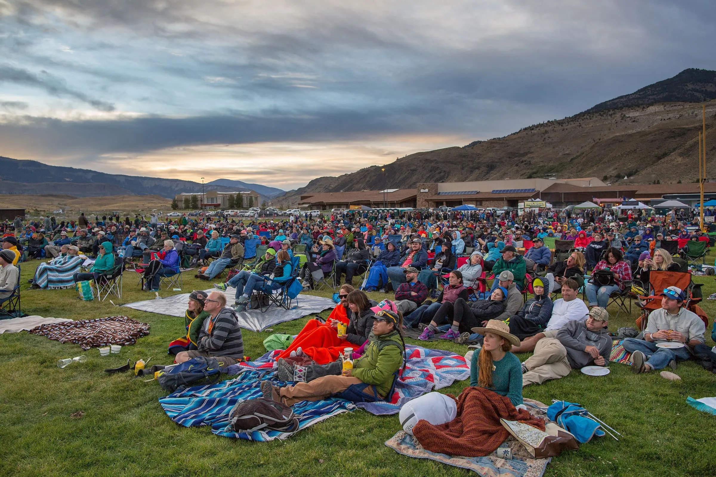 A large crowd sits on blankets and lawn chairs on a school football field below a mountain at dusk.