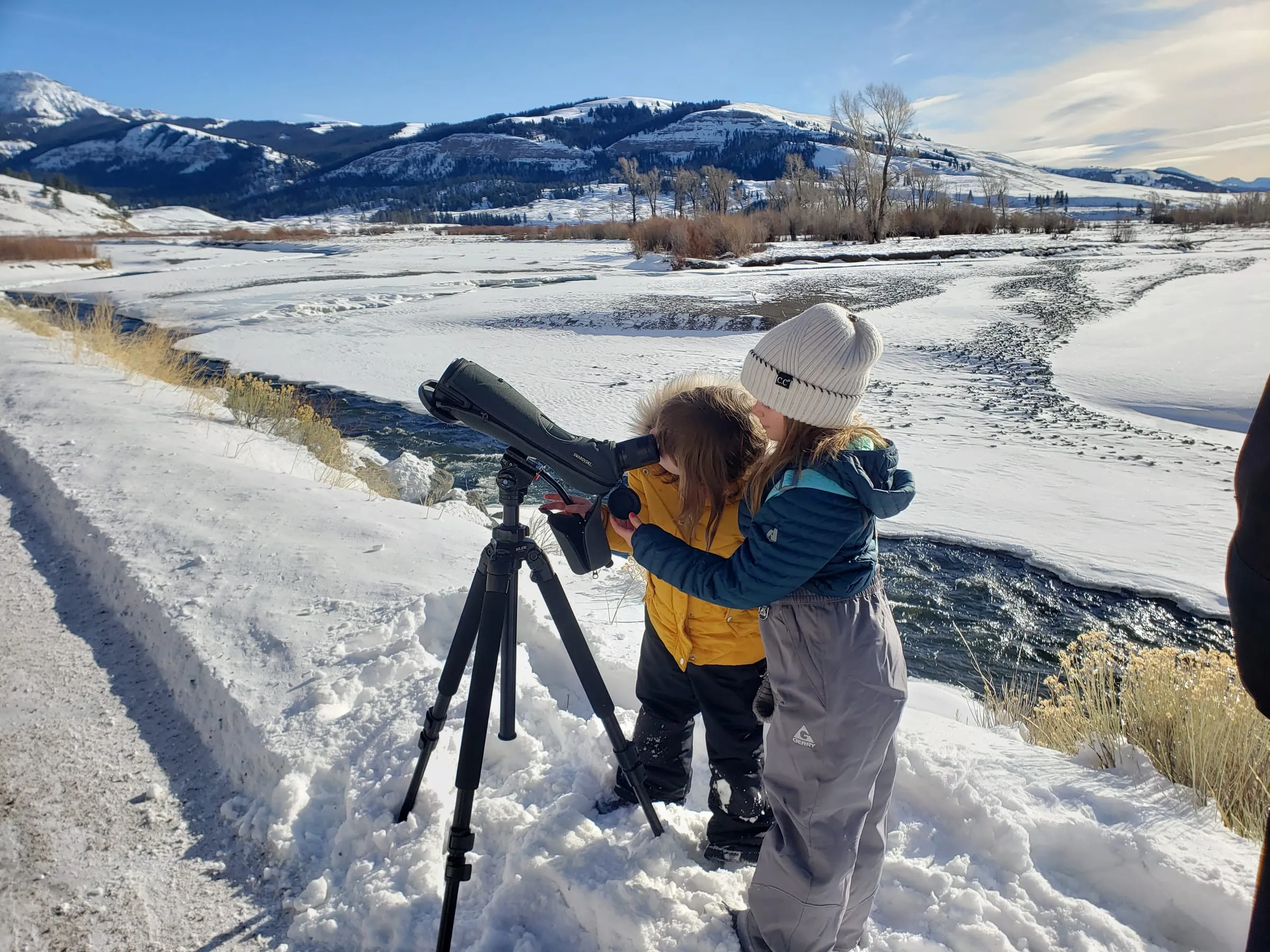 Two children in winter coats look through a spotting scope on a tripod above a snowy river valley.