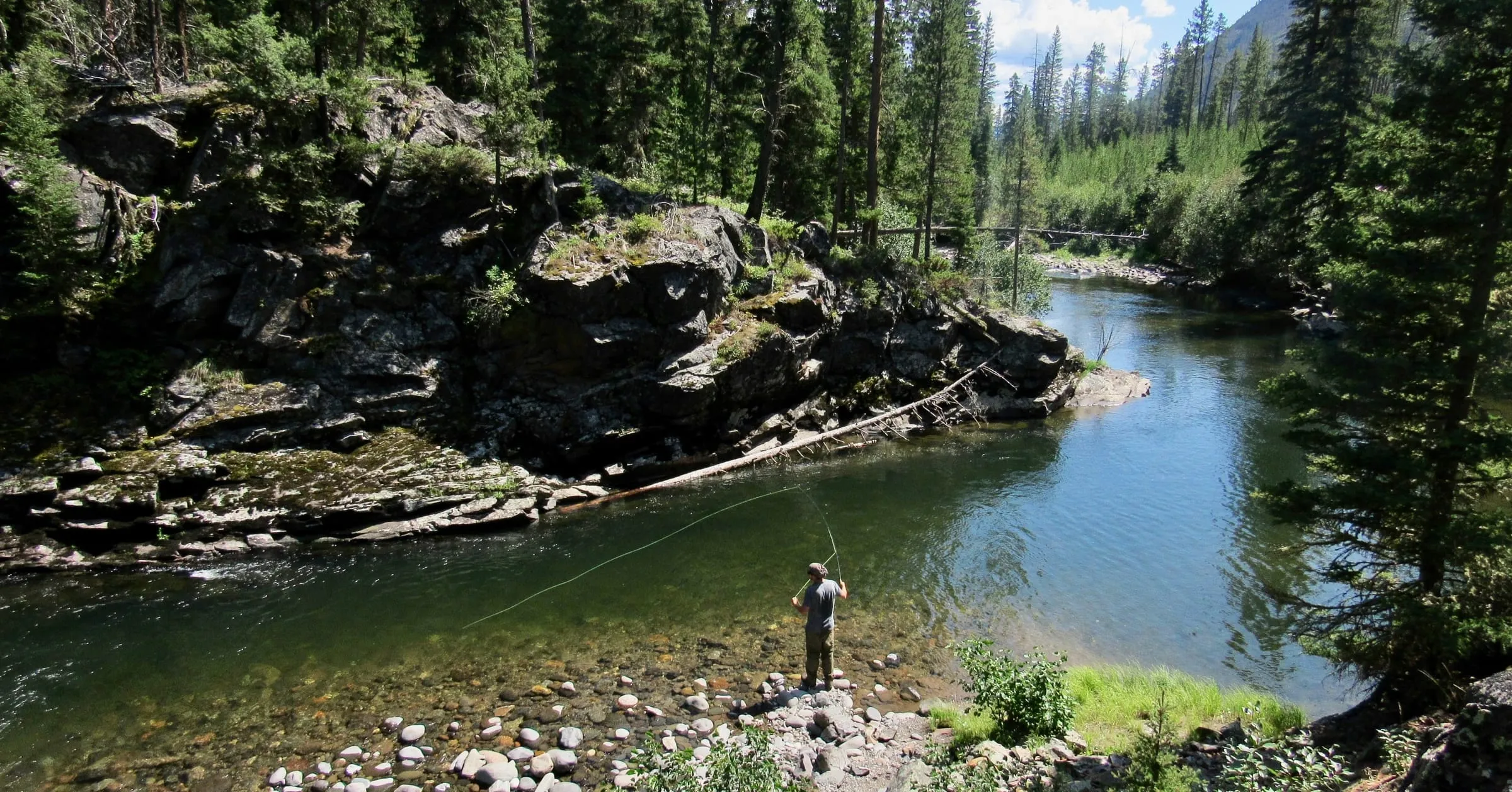 An angler casting a fly line into a clear green river pool below rocky ledges and conifers.