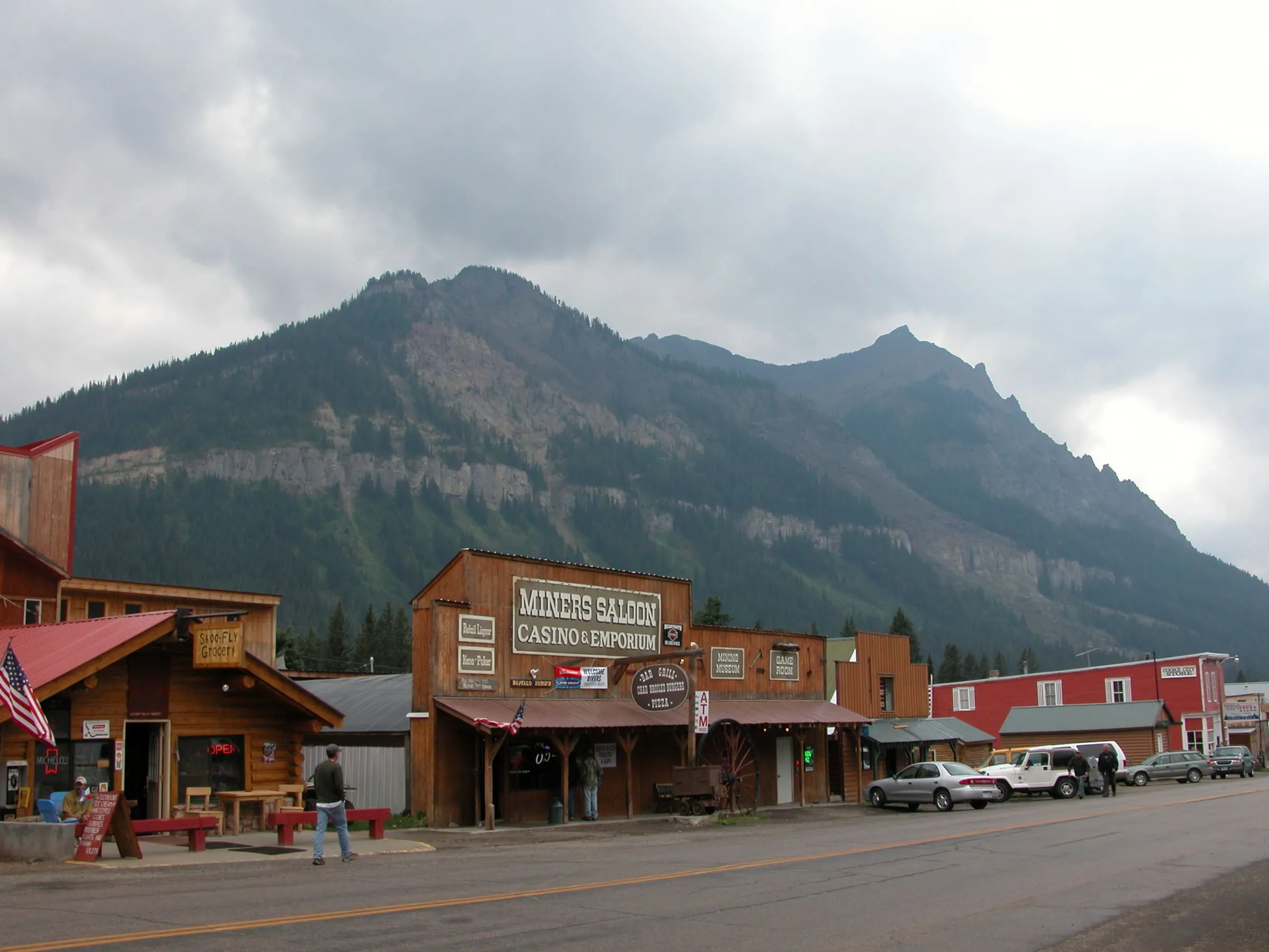 Wooden false-front buildings including the Miners Saloon line a street below a rugged mountain.