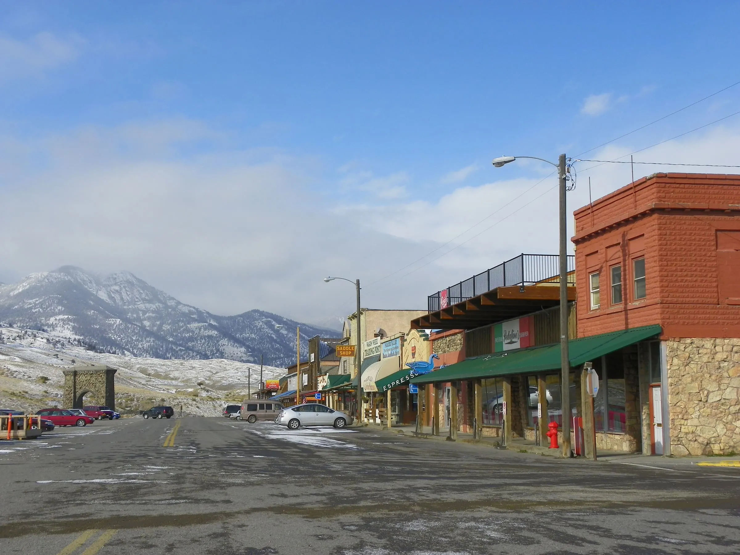 Park Street in Gardiner with a brick building and shop awnings, and the Roosevelt Arch and a snowy peak at the end of the street.