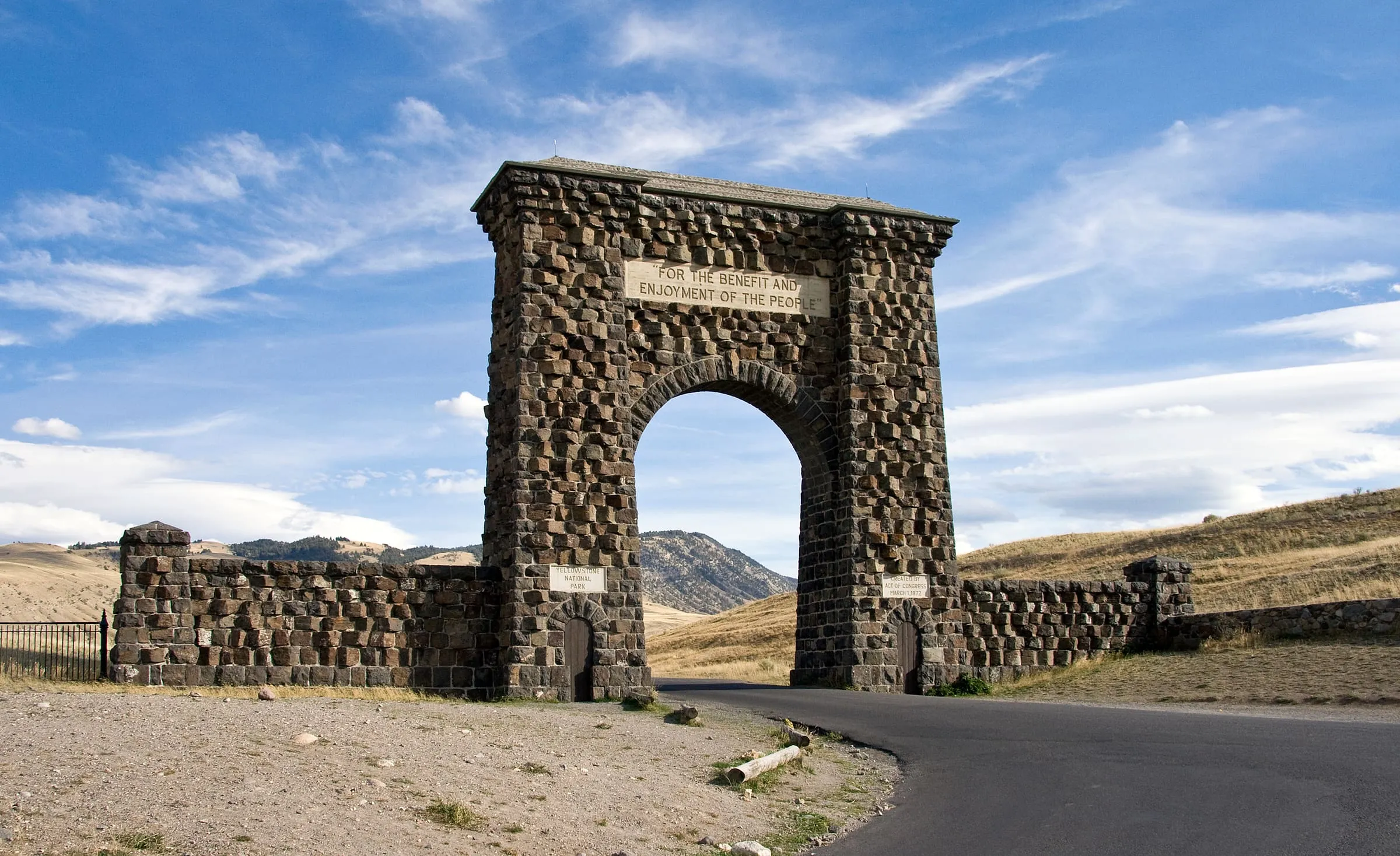 The stone Roosevelt Arch and its side walls stand beside a paved road under a blue sky with thin clouds.
